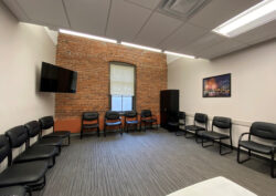 White room with brick accent wall on opposite end of photo. Black chairs around the outside of the room with a tv in the left corner