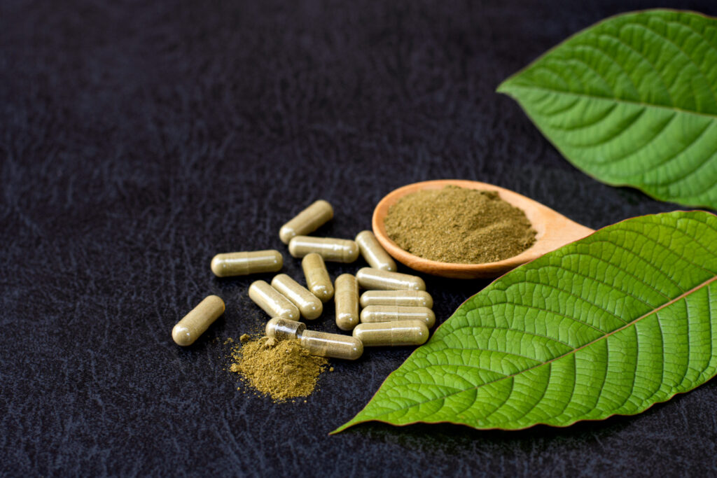 Kratom (Mitragyna speciosa) capsules with kratom powder and green leaves on wooden table background.