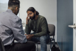 Over-the-shoulder shot of a doctor and man. The man looks sad and depressed after hearing news from the doctor. The man's eyes are closed and has one hand covering part of his face.