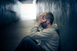 Profile view of a young homeless caucasian male - in his late 20s or early 30s - sitting in a dark, damp subway tunnel, his knees drawn up and his hands covering his face in desperation and despair. He is hungry, lonely and desperate. The man has short cropped hair and an unkempt beard. There is a look of sadness, loneliness and desperation in his eyes. He is sitting on a piece of old cardboard Horizontal image with room for copy space.