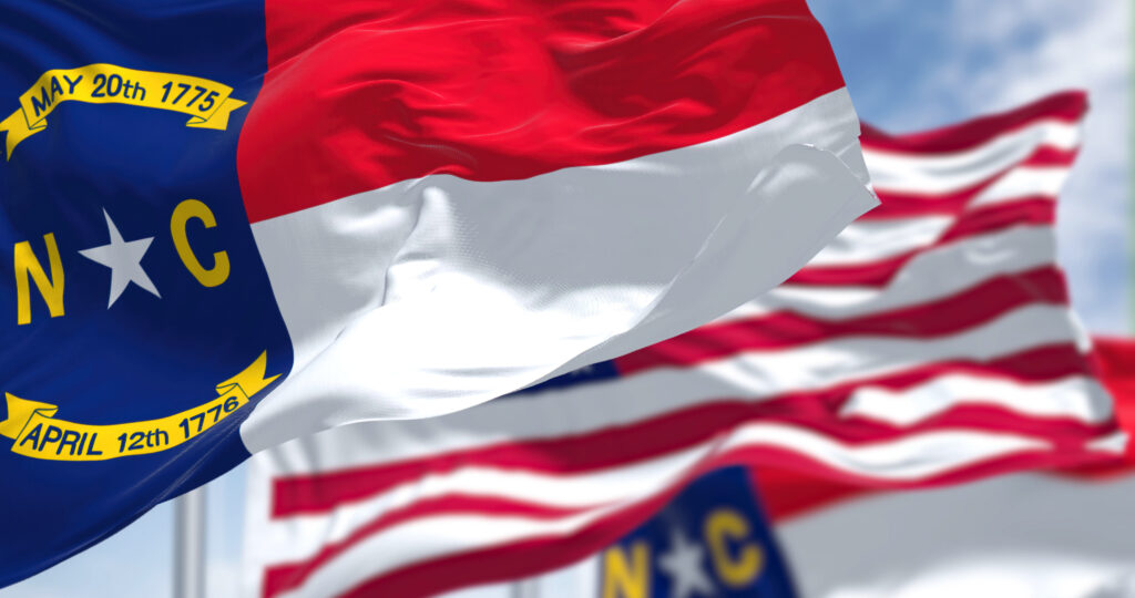 The North Carolina state flag waving along with the national flag of the United States of America. In the background there is a clear sky. North Carolina is a state in the Southeastern region of the United States