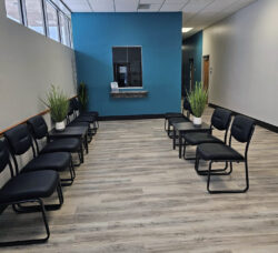 White room with blue accent wall. Waiting room with 2 rows of black chairs along the left wall and center of the room. Reception desk at the opposite end of the room on the blue wall